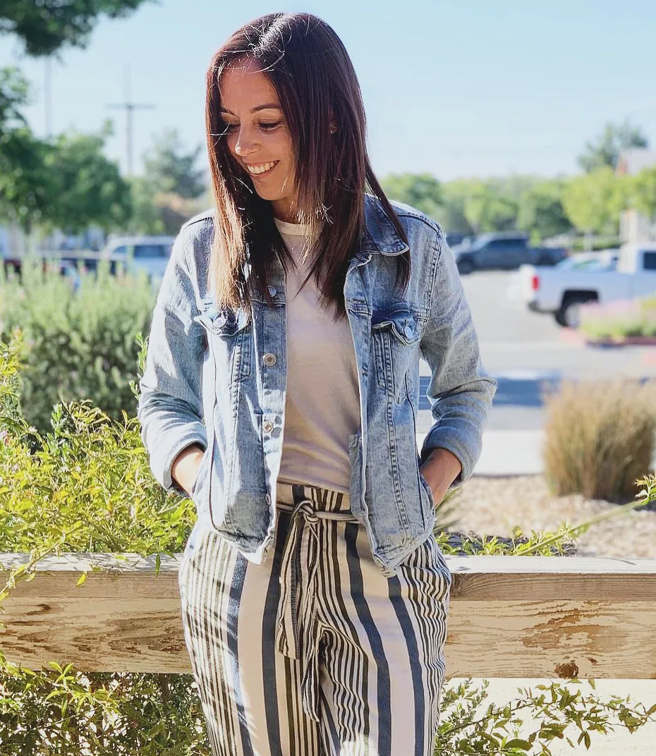 Ramey Krosky in a denim jacket, smiling and looking down, leaning on a wooden fence in the sun