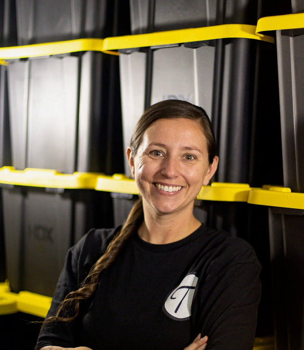 The owner of Tote-ably, arms folded and smiling, in front of shelves of black and yellow rental totes